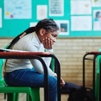 Menina negra, aparentando ter de 8 a 10 anos, de cabelos compridos, presos, está sentada em uma mesa escolar sozinha em uma sala. A mão segura o queixo com expressão chateada. Ela veste calça jeans e blusa verde