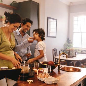 Uma família reunida na cozinha de sua sala. O pai oferece um cookie para o seu filho, enquanto a mãe ao lado acompanha a atividade familiar. A foto remete ao tema cookie caseiro