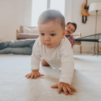 Bebê engatinhando no chão da sala de casa, com a supervisão do pai olhando do canto da sala