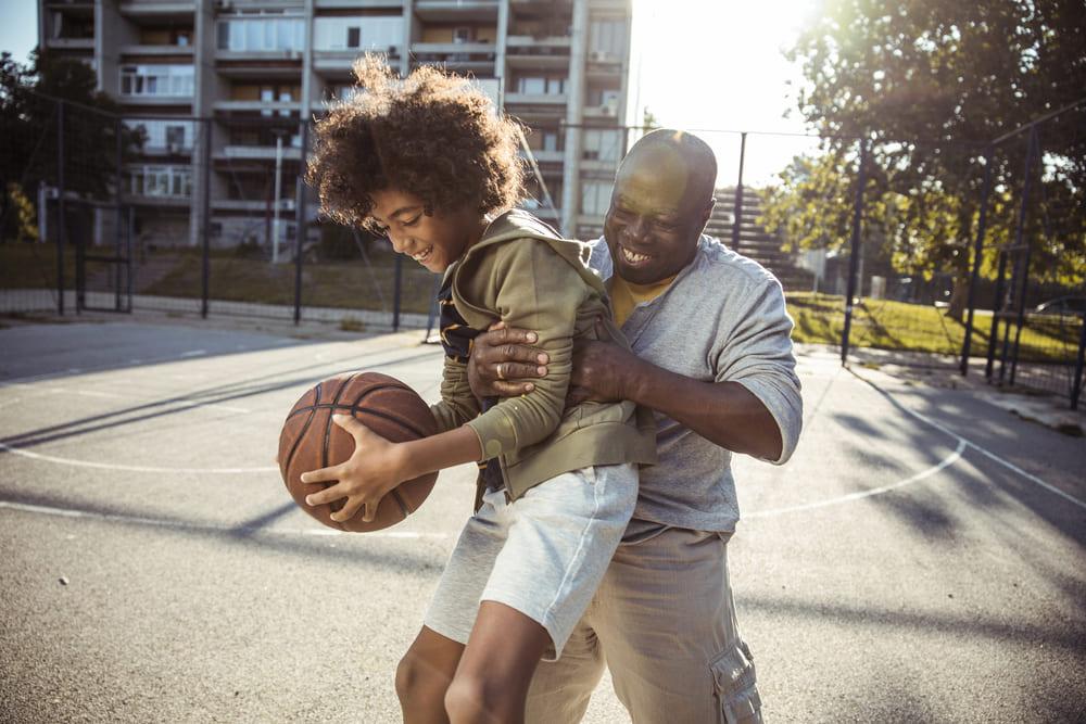 Na foto que representa os benefícios da atividade física, há um homem e um menino jogando basquete em uma quadra ensolarada. O homem está levantando o menino por trás, e, o menino está segurando a bola de basquete. Os dois estão vestidos com bermuda e casaco e estão sorrindo
