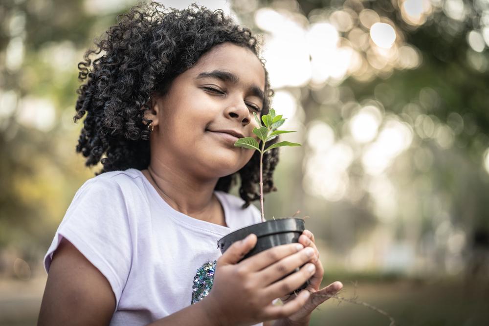 Uma menina segura um vaso pequeno com uma muda de planta entre as mãos. Ela está ao ar livre e de olhos fechados.