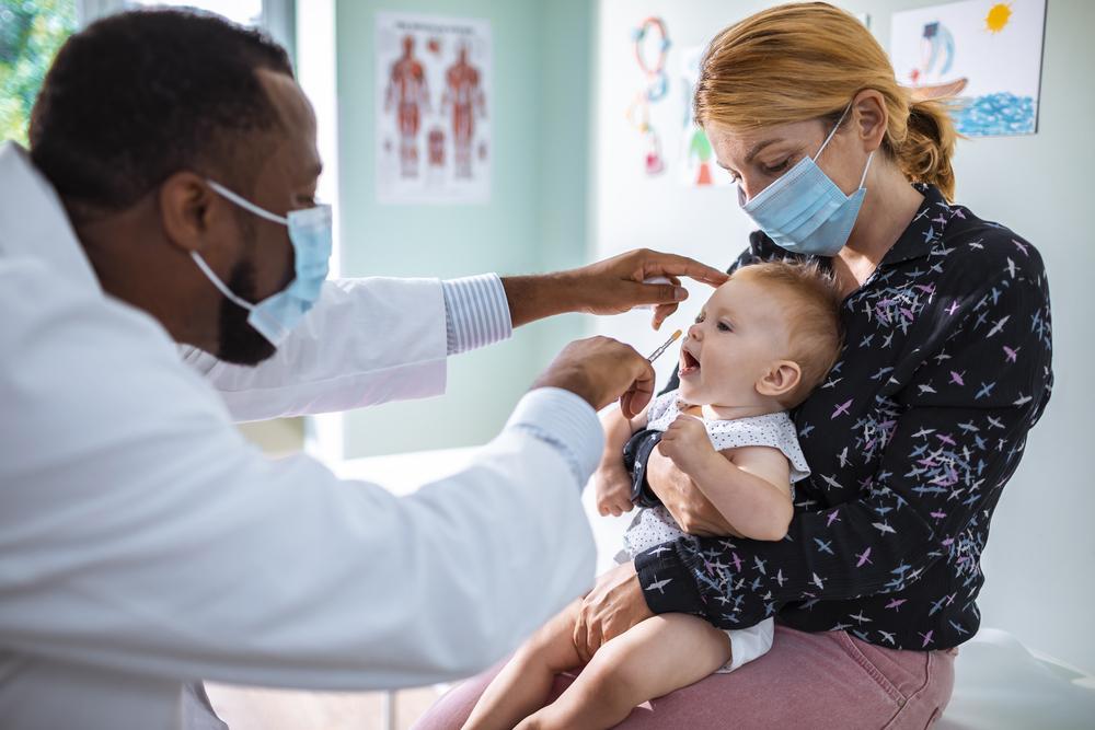  A masked doctor is holding a baby in his arms while the baby's mother looks on as the doctor administers a vaccine to the baby.