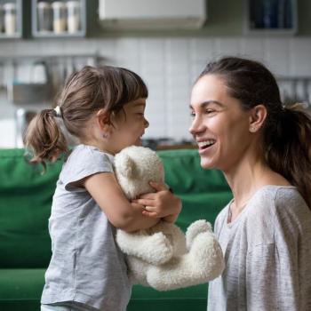 Mãe e filha se olham sorrindo, em frente a um sofá, na sala de casa. A mãe está abaixada no mesmo nível da menina, que abraça um ursinho de pelúcia.