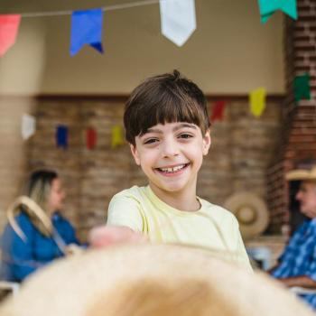 Criança sorrindo para a foto. Ao fundo, dois adultos conversam num cenário de festa junina, com bandeirinhas coloridas penduradas.