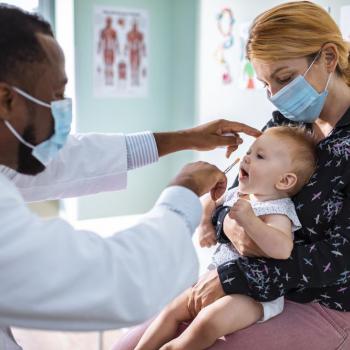 Em um consultório, um médico pediatra examina um bebê que está no colo de sua mãe. 