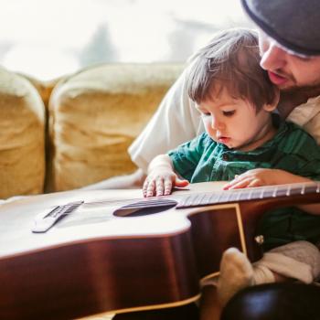 Um homem segura seu filho no colo e, juntos, eles seguram um violão, usando da música para estimular o desenvolvimento infantil.