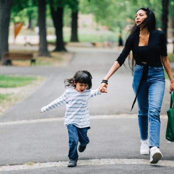 Uma mãe lida com a birra infantil em um parque. De mãos dadas com sua filha, a criança chora e está com cara de brava. 