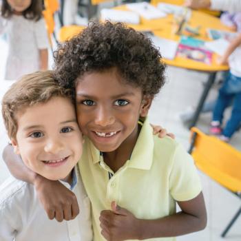 A foto mostra dois meninos de cores e aparências distintas sorrindo, se abraçando e olhando para a câmera, dentro de uma sala de aula. 