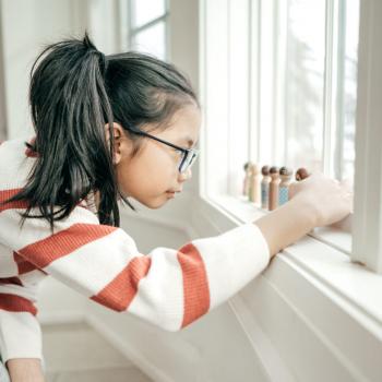 Na imagem, há uma menina vestida com blusa listrada e calça cinza, usando rabo de cavalo e óculos. Ela está agachada em frente à janela, enfileirando suas mini bonecas de madeira demonstrando perfeccionismo, uma das características do autismo 