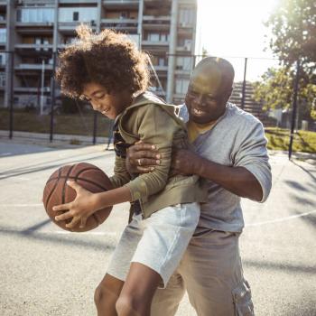 Na foto que representa os benefícios da atividade física, há um homem e um menino jogando basquete em uma quadra ensolarada. O homem está levantando o menino por trás, e, o menino está segurando a bola de basquete. Os dois estão vestidos com bermuda e casaco e estão sorrindo