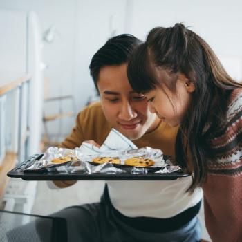 Um pai e sua filha estão em frente a um forno segurando uma bandeija de cookies na cozinha, como exemplo de atividades de dia dos pais. 