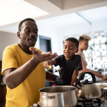 Pai e filho cozinham juntos como uma homenagem de dia dos pais. O pai está mexendo uma panela e o filho, ao seu lado, o ajuda. 