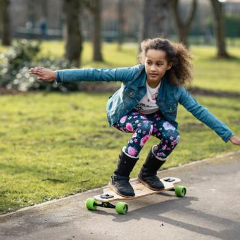 Foto de uma menina andando de skate ao ar livre, em um lugar plano. Um incentivo a pratica de skate infantil.