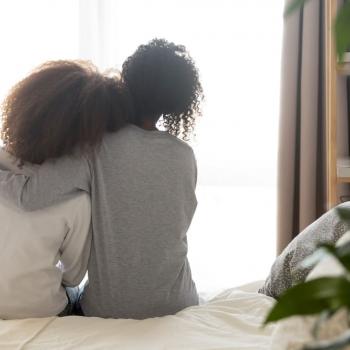 Mãe e filha se abraçam sentadas na cama.