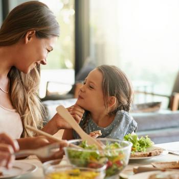 Na imagem, há uma mulher e uma menina sentadas em frente à mesa de almoço, em uma sala de estar. A mulher está olhando carinhosamente para a menina, que, por sua vez, está sorrindo para ela. Nos pratos, há alface com legumes, fatias de pão integral e um suco de laranja