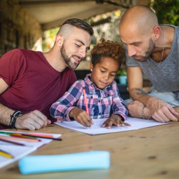 Dois homens observam sorrindo a filha desenhar em uma folha de papel, em uma mesa com lápis de cor espalhados e papéis.