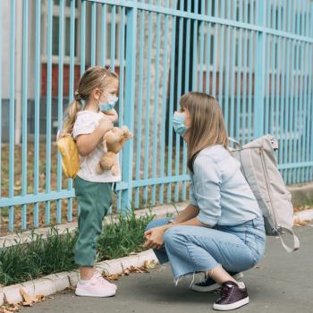 Uma mãe está agachada em frente a sua filha, perto de um portão. A menina está de mochila, olhando para a mãe e segurando um urso de pelúcia.