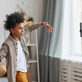 Um menino está fazendo uma pose de braços abertos em frente ao celular, que está apoiado em um tripé com ring light. 