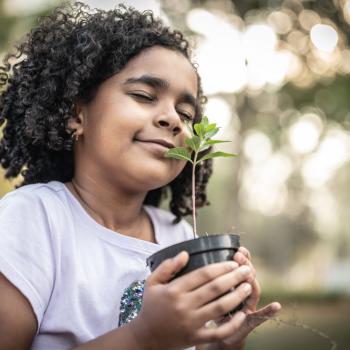 Uma menina segura um vaso pequeno com uma muda de planta entre as mãos. Ela está ao ar livre e de olhos fechados.