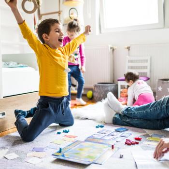 Sala com um adulto, onde só é possível ver suas pernas usando calça jeans e meia. Na mesma sala, estão três crianças. Uma garota com cerca de 1 ano de idade em segundo plano, branca, de cabelos curtos, de costas brincando com uma boneca. Outra garota com cerca de quatro anos, branca, cabelos loiros presos, calça jeans e blusa de manga comprida. Ela parece estar feliz dançando. A criança em destaque é um menino, branco, com cabelos curtos, de braços abertos comemorando uma vitória em um jogo 