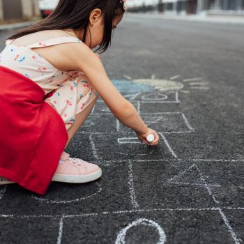 Uma menina de vestido e tênis está agachada na rua desenhando uma amarelinha no chão. A foto faz alusão ao tema de brincadeiras de rua