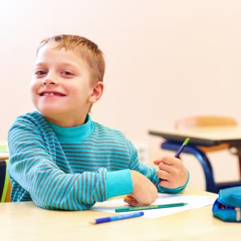 Criança aparentando ter 7 anos de idade, branca, loira, veste camiseta de manga longa azul sentado na classe da escola. Na mesa escolar, materiais para desenho como lápis e papel. 