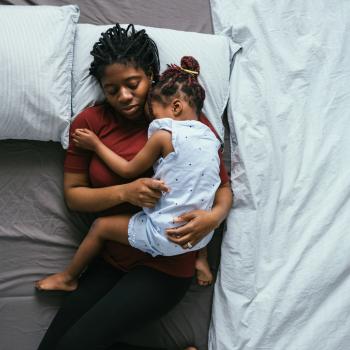 Foto de uma mãe solo dormindo com a filha no colo em sua cama. As duas estão no meio da cama que está com as laterais vazias