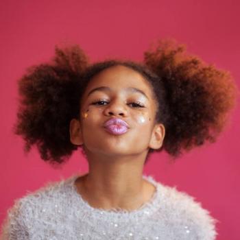 Foto de uma menina usando maquiagem colorida e uma blusa felpuda fazendo bico com a boca sobre fundo rosa. 