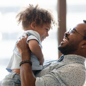 Uma criança é carregada pelo seu pai. Os dois estão brincando e sorrindo. A imagem remete ao tema crise dos três anos