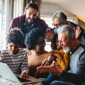 Imagem de uma família reunida na sala, olhando com felicidade a tela de um notebook. Ela é composta por um casal jovem e duas crianças, além de outros dois membros que são os avós dessas crianças. A foto remete ao tema rede de apoio materno