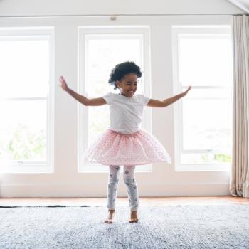 Menina vestindo um tutu de balé no meio da sala de casa. Ela se encontra com um semblante feliz e dançando pelo espaço. A foto faz alusão ao tema ballet infantil.