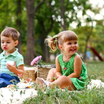 Um menino e uma menina estão sentados em uma toalha em um parque. Sobre ela temos algumas cestas e lanchinhos. O cenário todo remete muito a um dia de piquenique