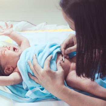 Imagem de um bebê deitado, envolvido em uma manta azul, chorando, enquanto a cuidadora limpa seu coto umbilical com um cotonete