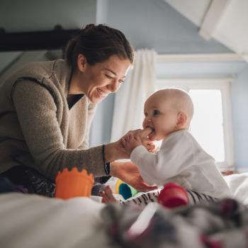 Um bebê está sentado na cama, mordendo o dedo da sua mãe que está na sua boca. A imagem remete ao tema de cárie formada pelo uso da mamadeira