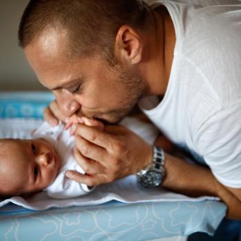 Bebê deitado na cama de barriga para cima com seu pai o acariciando e beijando nos seus pezinhos