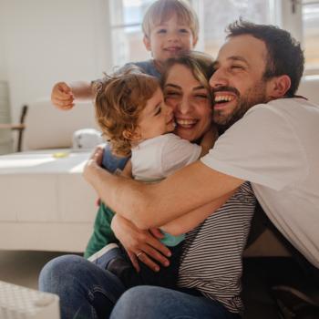 Família composta por pai, mãe e dois filhos de abraçando sentados no chão em frente ao sofá da sala