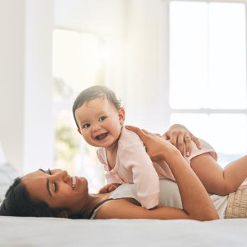 Mãe morena de cabelo preto liso deitada na cama de barriga para cima segurando seu bebê 