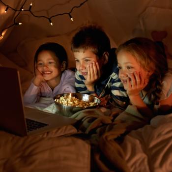 Três crianças, sendo um menino e duas meninas, deitados assistindo no notebook enquanto comem pipoca. Ao fundo, luminárias em uma cabana.