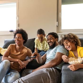 Imagem de uma família assistindo a filmes infantis. Mãe e pai estão sentados no chão e os filhos no sofá. Todos estão gargalhando.