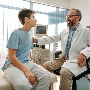 Imagem de um ambiente hospitalar em que um médico tranquiliza um paciente menino, ao colocar a mão no ombro dele. O médico está de jaleco branco, óculos e tem barba e o menino está vestindo uma camiseta azul