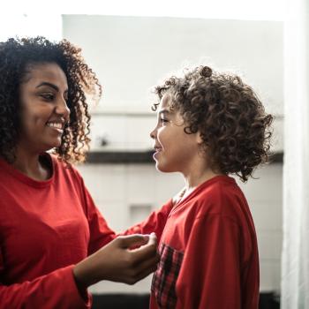 Mulher sorrindo com seu filho, ambos usando um pijama vermelho e conversando. Ao fundo, um quarto.