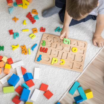 Em um piso de madeira com um tapete cinza, está sentado um menino,vestindo uma camiseta branca. Ele está brincando com um tabuleiro de encaixar letras, enquanto segurando uma delas. Há letras e outras peças coloridas espalhadas ao seu redor.