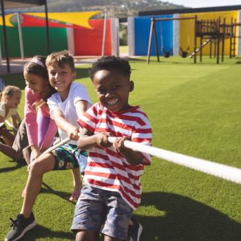 Em um parque ao ar livre, três meninos e duas meninas estão se divertindo em uma brincadeira de cabo de guerra. As crianças, sorridentes, estão puxando a corda.