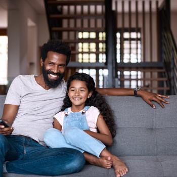 Em uma sala, um homem, vestindo uma camisa cinza, e uma menina, vestindo uma jardineira jeans e uma camisa branca, estão sentados em um sofá cinza. O homem está segurando um controle remoto, apontado para frente. O homem e a menina estão sorrindo. 