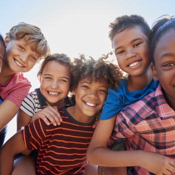 Ao ar livre, em um dia ensolarado, seis crianças - três meninas e três meninos - estão abraçadas entre si, sorrindo enquanto brincam.