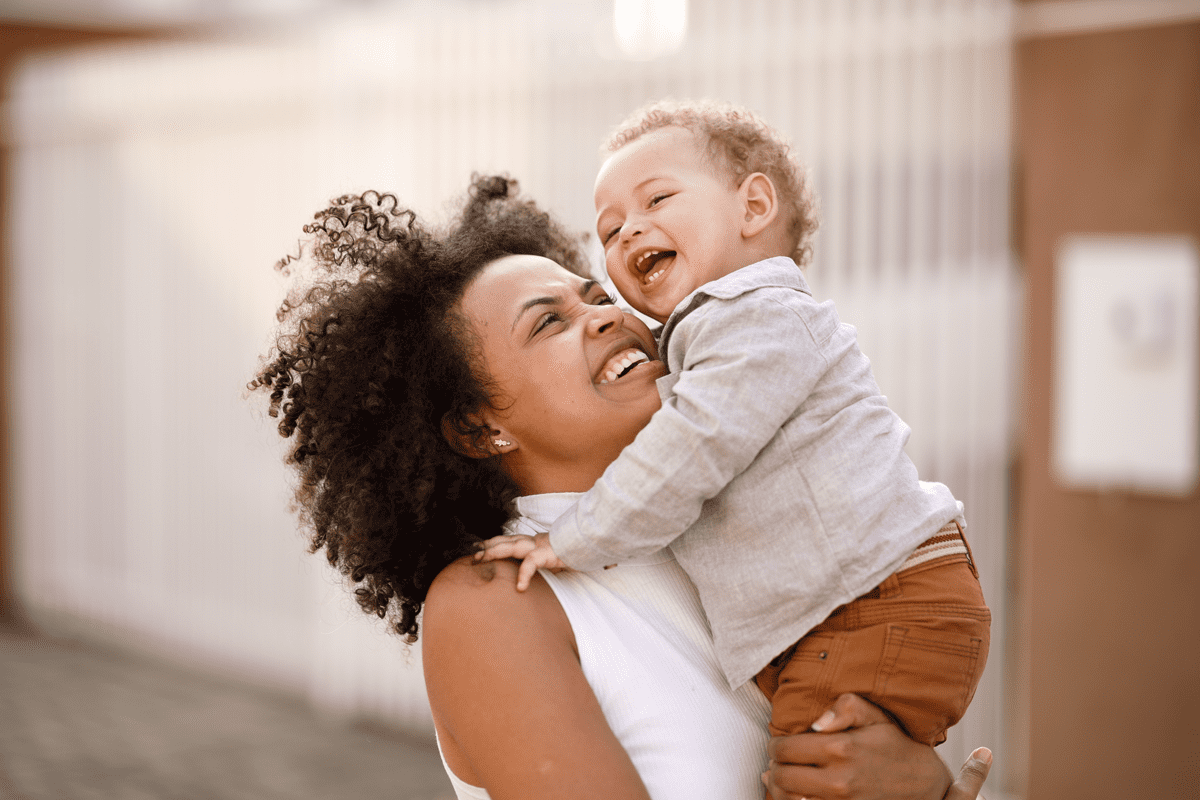 Mãe sorridente carrega o filho no colo, ilustrando a fase conhecida como terrible two. 