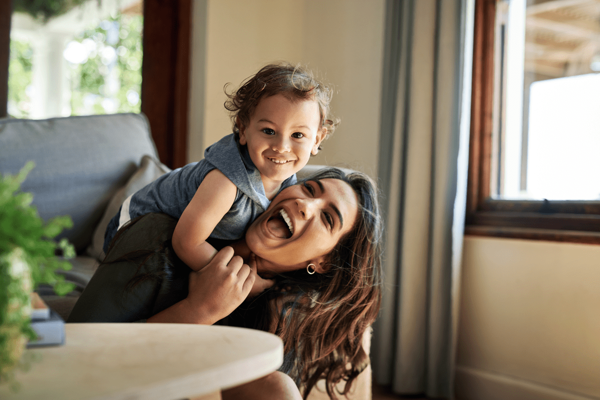 Uma criança de 3 anos está apoiada nas costas de sua mãe. Ambos estão na sala de estar e sorriem.