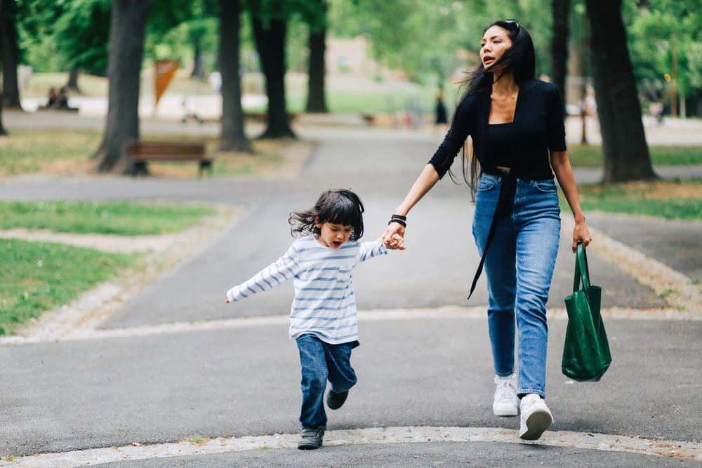 Uma mãe lida com a birra infantil em um parque. De mãos dadas com sua filha, a criança chora e está com cara de brava. 