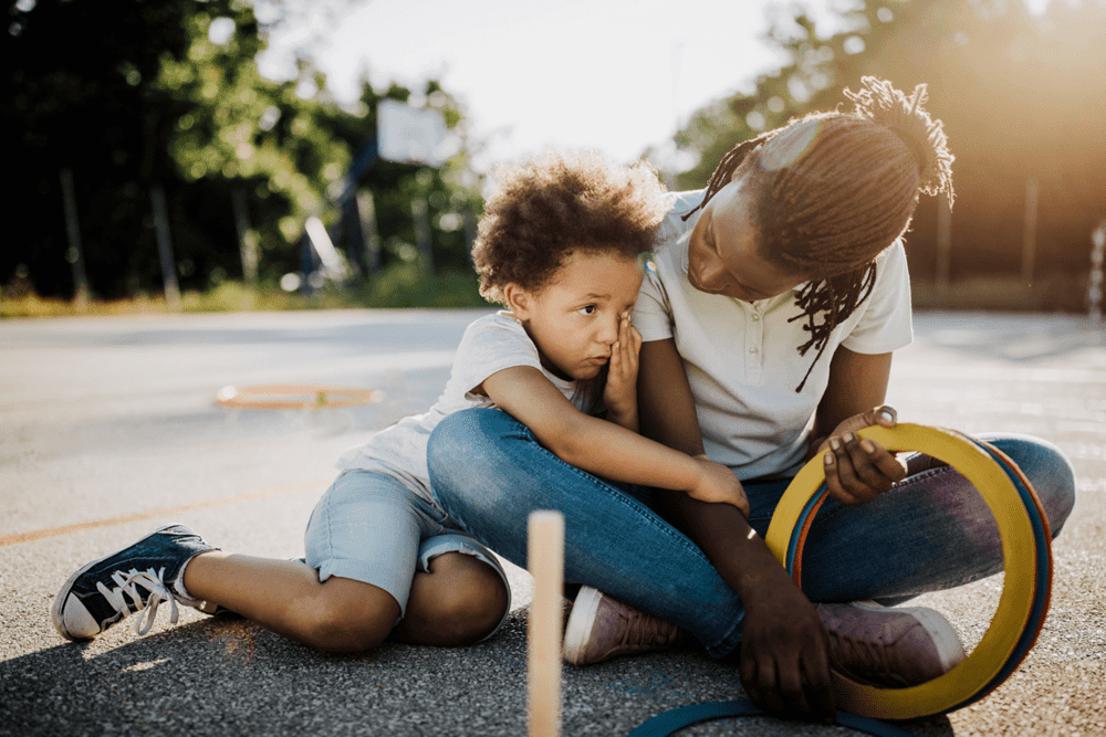 Mãe e filho, uma criança de 6 anos, estão sentados no chão de um espaço ao ar livre. A mãe olha para o filho, que o abraça.