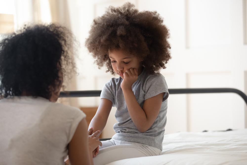 Para ilustrar a depressão infantil na foto, mãe consola a filha, que está sentada, com expressão triste, em cima da cama.